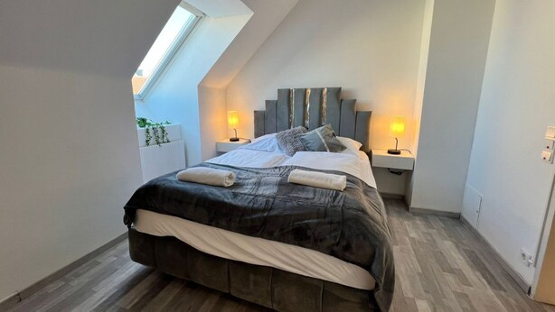 Ein Schlafzimmer mit schrägen Wänden, einem Bett mit dunklem Kopfteil, zwei Nachttischlampen und einem Dachfenster.