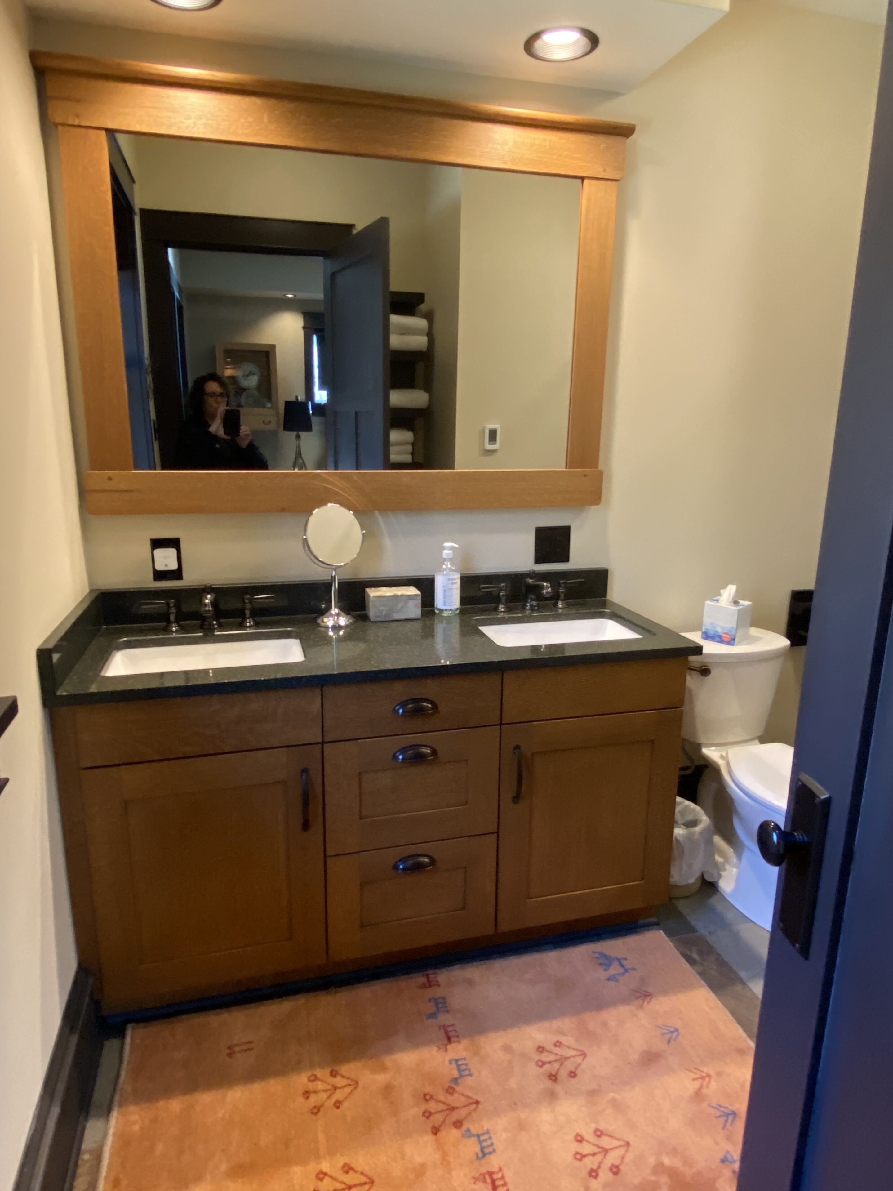 Ein Badezimmer mit einem Holzkosmetikschrank, zwei Waschbecken, einem Spiegel, einer Toilette und einem Teppich mit Muster.