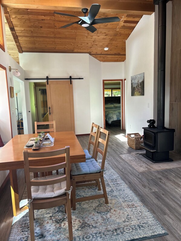 Un coin repas avec une table et des chaises en bois, un ventilateur de plafond, un poêle noir et une vue sur un espace extérieur verdoyant.
