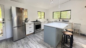 A modern kitchen with a stainless steel refrigerator, double oven, and a large island with stools. - Huckleberry Hideaway - an idyllic off-grid cabin on Savary Island, BC. Canada. (Lund)