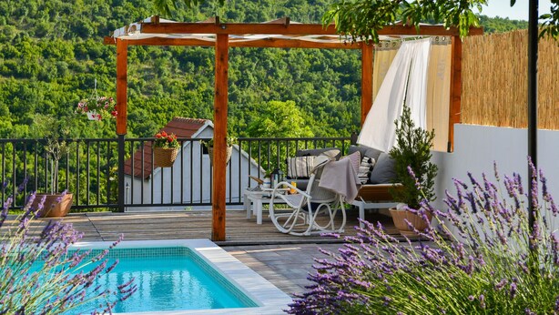 Una terraza de madera con piscina, mobiliario exterior y una pérgola con toldo blanco.