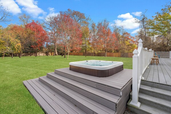 A hot tub on a wooden deck with steps leading up to it, surrounded by a grassy yard and trees with autumn foliage.