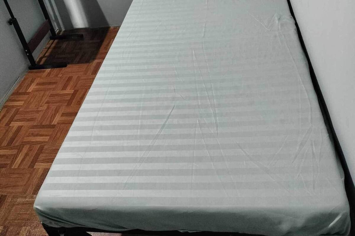 A small, single-bed room with a wooden floor, a metal desk, and a grey mattress.