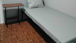 A small, single-bed room with a wooden floor, a simple desk, and a plain white wall.