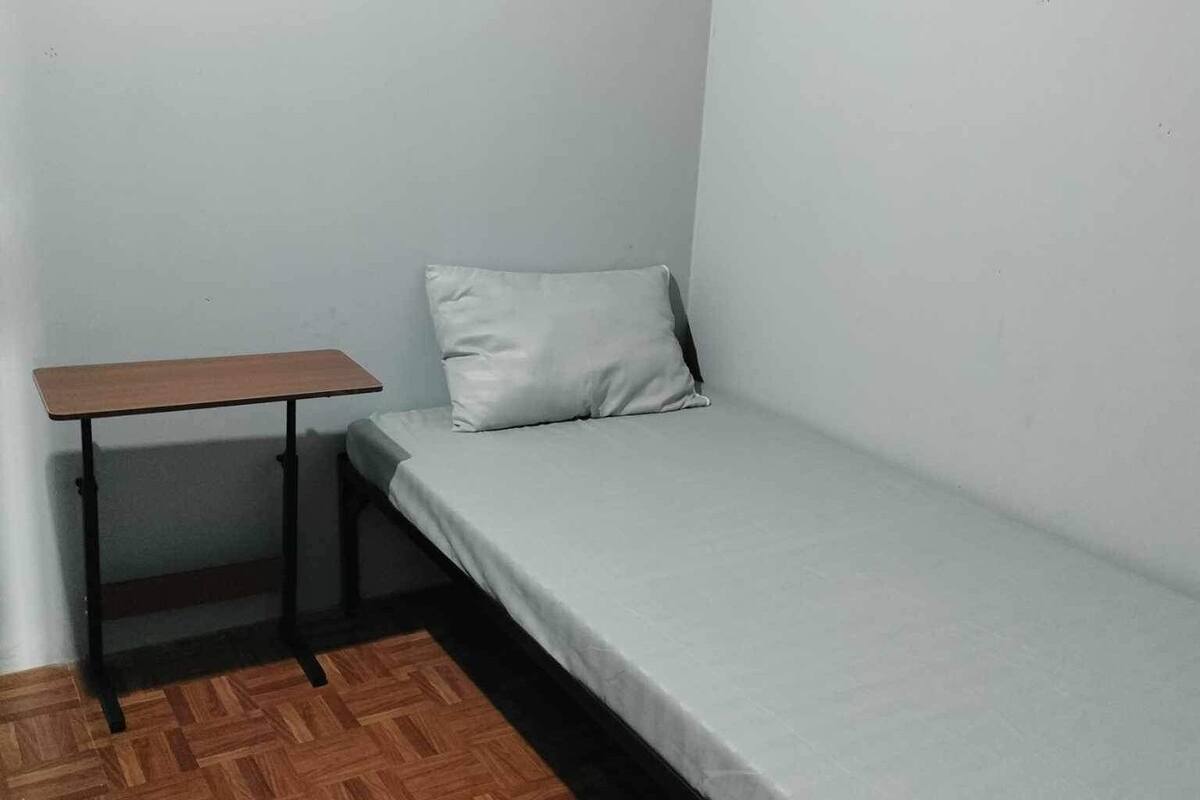 A small, single-bed room with a wooden floor, a simple desk, and a plain white wall.