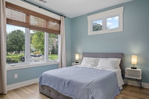 Un dormitorio con una cama, dos mesitas de noche, una ventana con persianas y vistas al exterior.