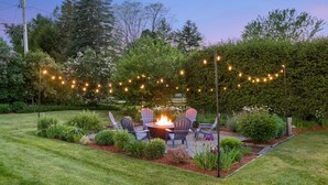 Un jardín bien cuidado con una fogata, asientos exteriores y luces colgantes.