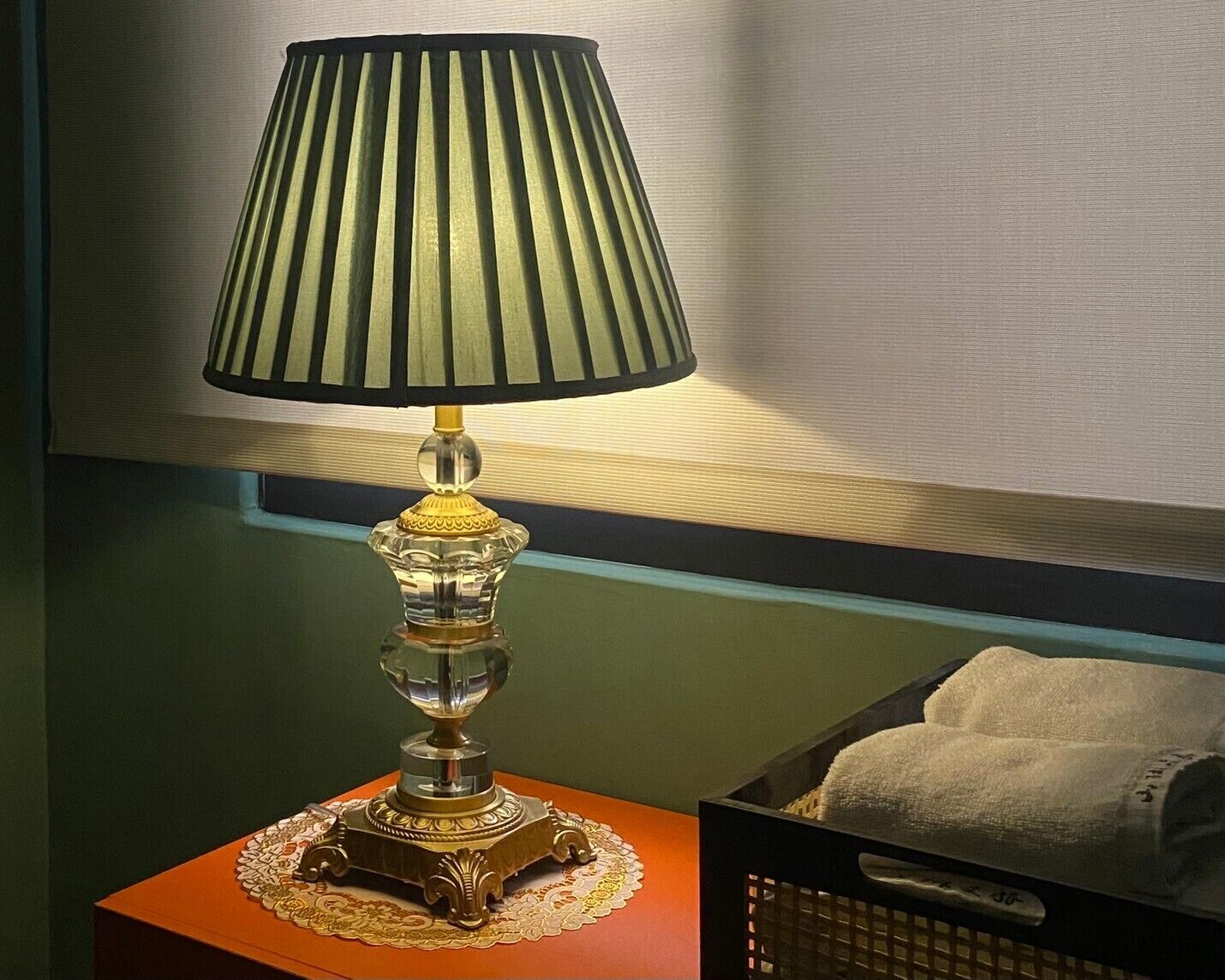 A table lamp with a pleated lampshade and a decorative base, placed on a wooden table with a lace doily.