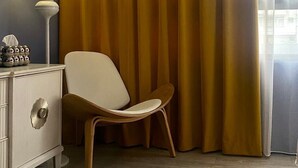 Um quarto com penteadeira branca, luminária, cadeira e uma janela grande com cortinas.