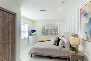 A bedroom with a bed, a TV, a desk, and a window with blinds.