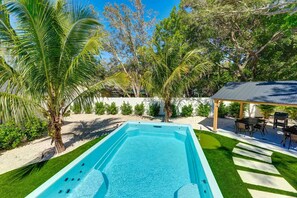A rectangular swimming pool with a wooden deck surrounding it, a covered outdoor seating area, and a variety of tropical plants.