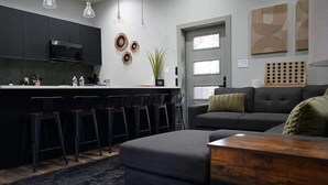 A modern kitchen with dark cabinets, a central island with bar stools, a grey sofa, and a wooden side table.