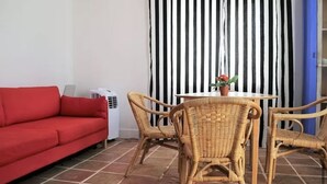 Ein Wohnbereich mit einem roten Sofa, Korbstühlen, einem kleinen Tisch mit Topfpflanze und einer weißen Klimaanlage.