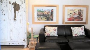Ein Wohnzimmer mit einer schwarzen Ledersofa, zwei passenden Hocker, einer weißen Tür mit abblätternder Farbe und zwei gerahmten Bildern an der Wand.