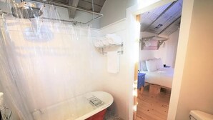 Ein Badezimmer mit einer roten Badewanne auf Pfotenfüßen, einem weißen Waschbecken und einer Toilette. Ein Duschvorhang und ein Regal mit Handtüchern sind vorhanden.
