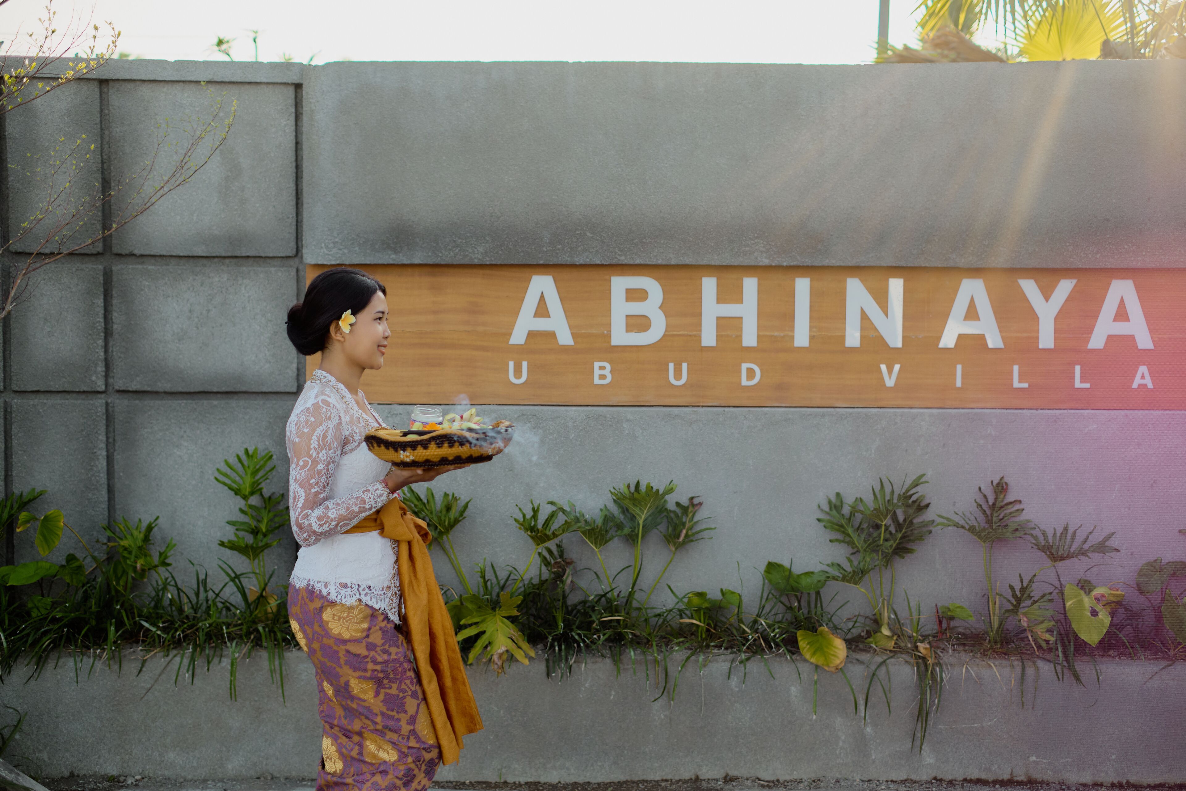 A woman carrying a tray with food in front of a sign that reads "ABHINAYA Ubud Villa."