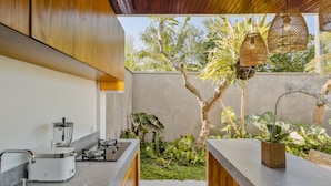 A modern kitchen with a built-in stove, a blender, and a sink. There is a small garden area with plants and a tree visible through the window. - Abhinaya Ubud Villa (Ubud)