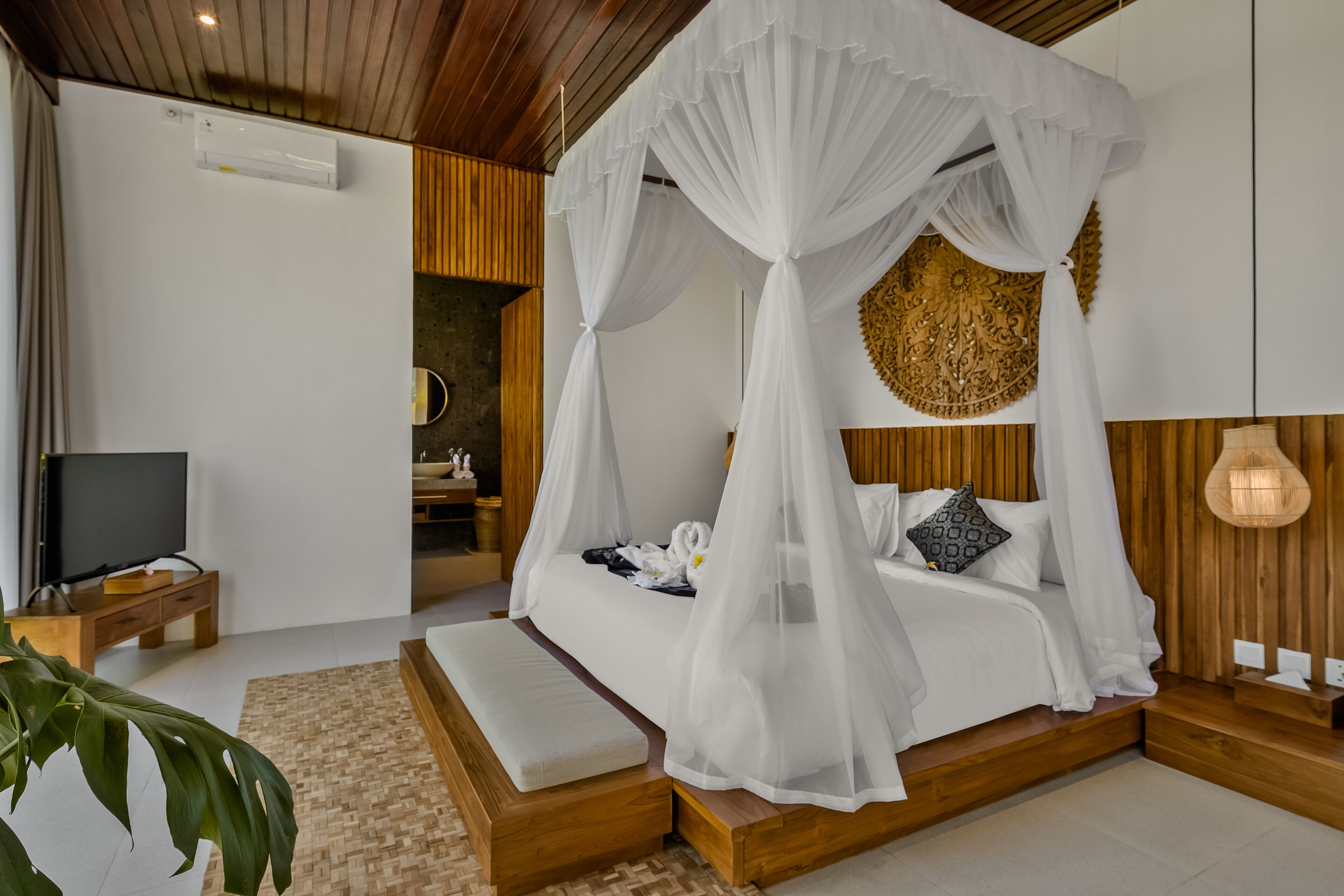 A modern bedroom with a four-poster bed, a television on a wooden nightstand, and a large plant in the corner.