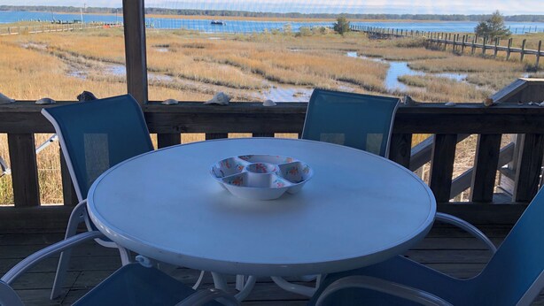 A round table with a white tablecloth and a bowl, surrounded by blue chairs on a wooden deck overlooking a beach with a wooden fence and a body of water in the background. - Riptide’s Crossing:  A waterfront getaway w/deep water dock and incredible views (Chincoteague)