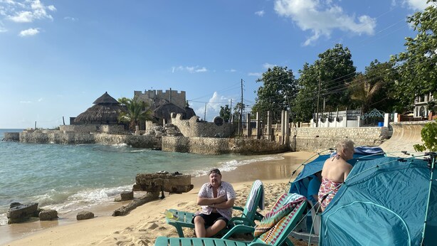 A beach with a stone wall and a thatched-roof building in the background. There are two people sitting on green lounge chairs under a blue tent. - The Belair SeaSide Villas. 4 suites, 2 guests per Suite. 1, 2/bedroom/3 guests (Belmont)