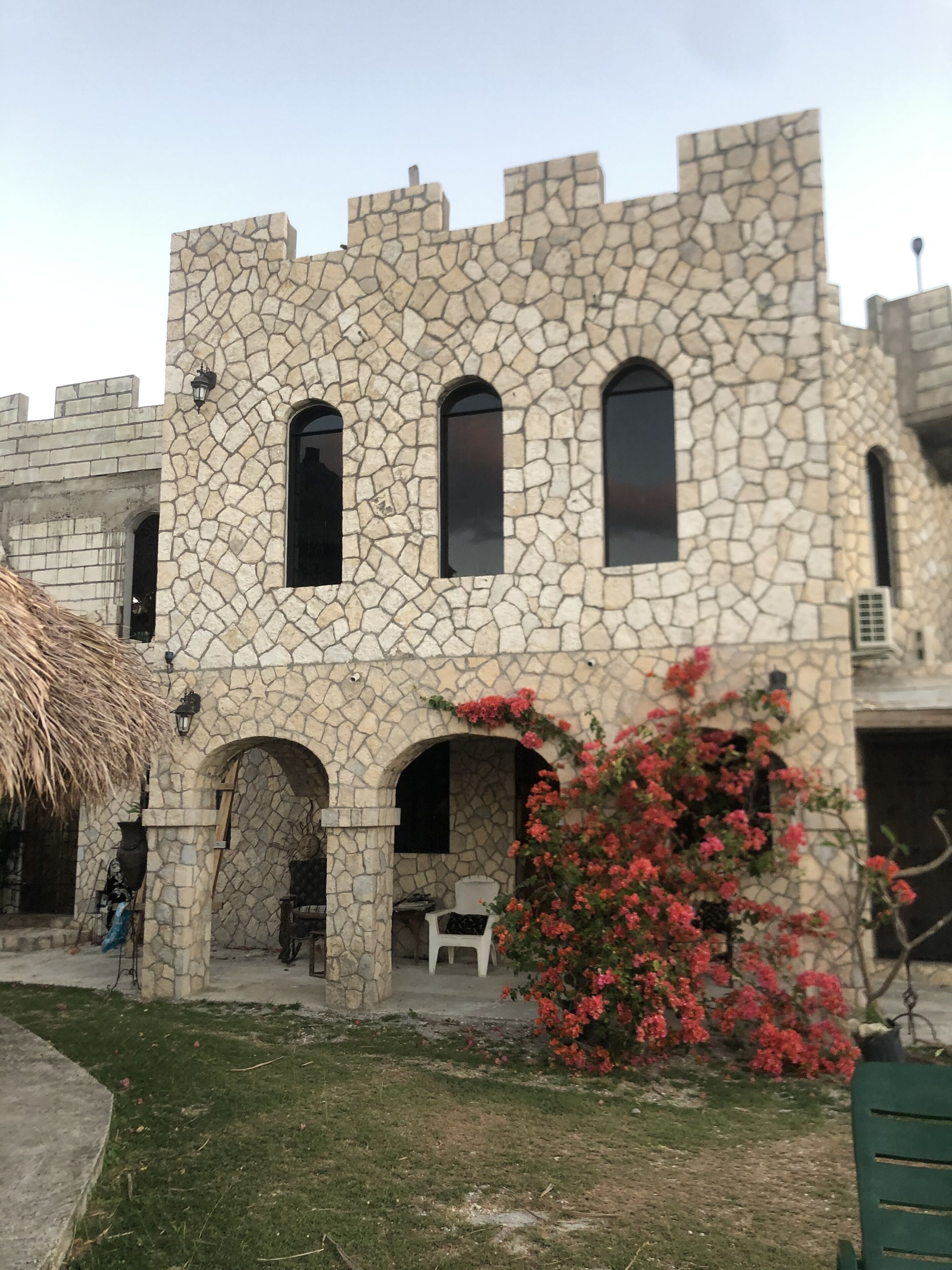 Een stenen gebouw met boogvormige deuropeningen en ramen, een rieten dak en een kleine tuin met rode bloemen.