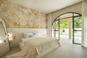 A modern bedroom with a stone wall, a large bed, and a view of a pool and greenery.