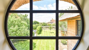 Une fenêtre circulaire donnant sur un jardin bien entretenu et une maison traditionnelle à toit de chaume.