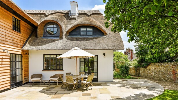 Une maison avec un toit de chaume, une terrasse avec une table et des chaises, et une pelouse bien entretenue.