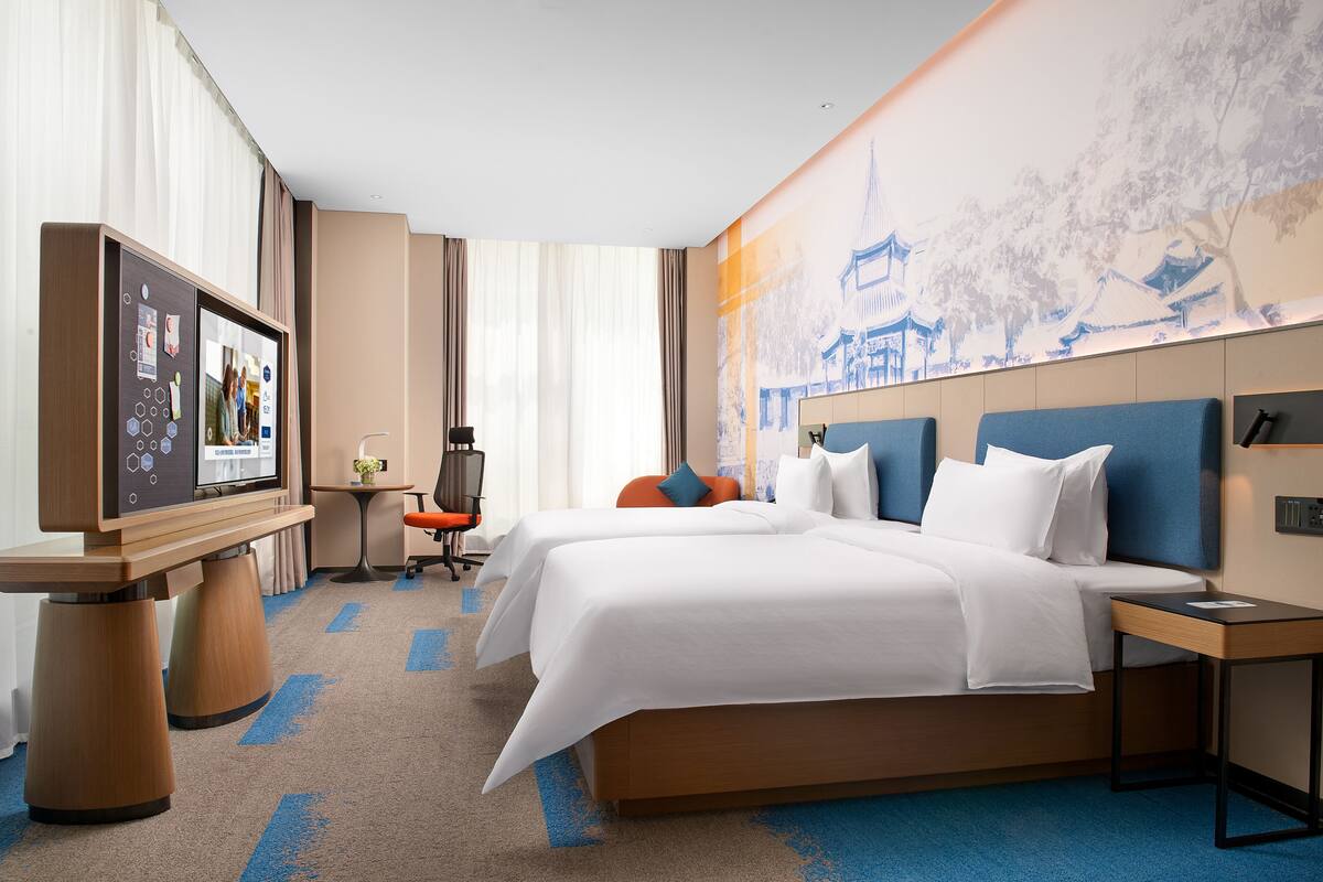 Hotellihuone, jossa on kaksi sänkyä, työpöytä tietokoneella, televisio ja seinä, joka on koristeltu perinteisellä seinämaalauksella.