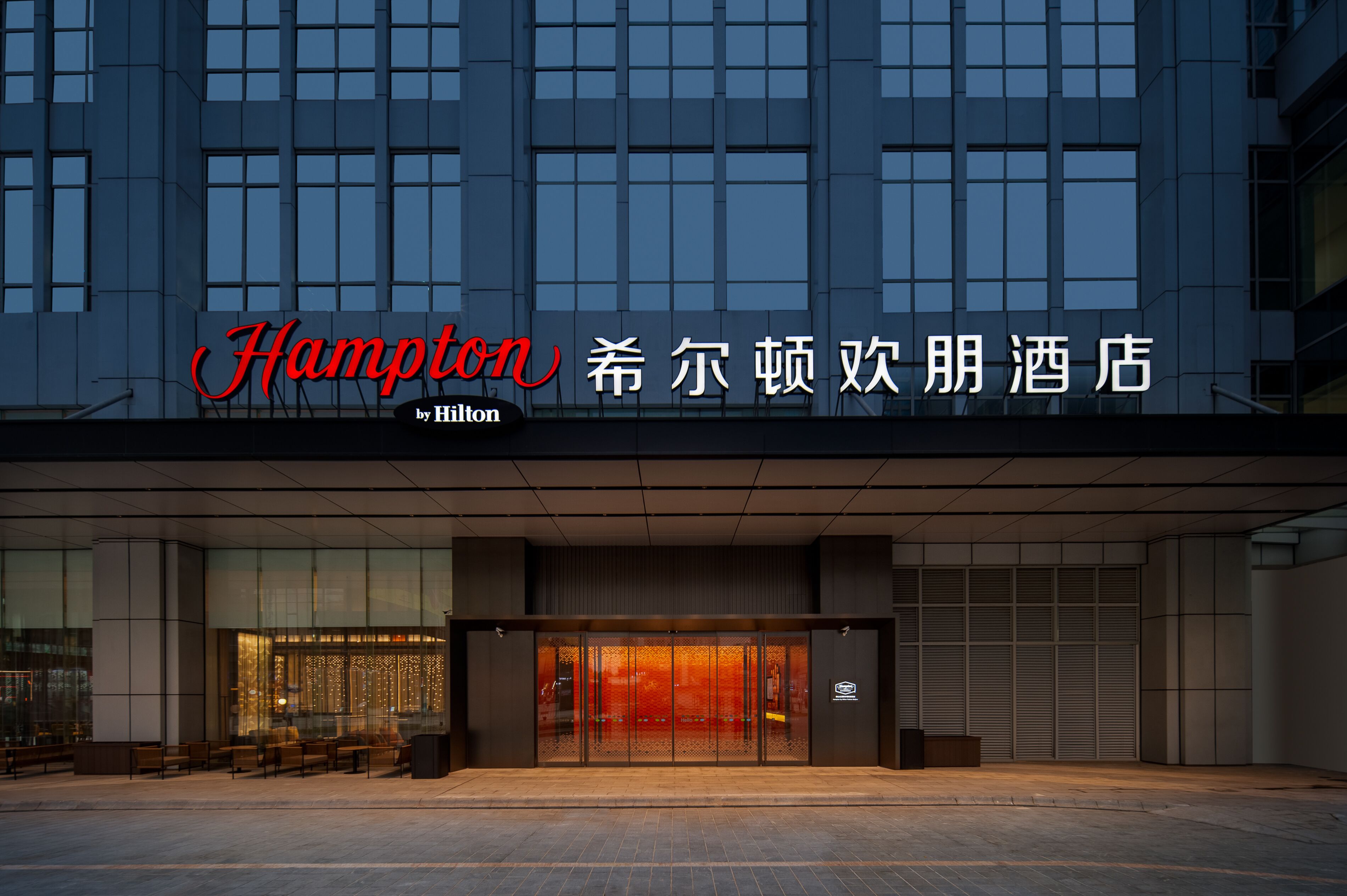 Une entrée d’hôtel moderne avec une façade en verre, un espace extérieur aménagé avec des sièges, et une enseigne portant les noms « Hampton » et « Hilton ».