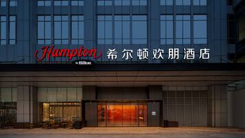 Une entrée d’hôtel moderne avec une façade en verre, un espace extérieur aménagé avec des sièges, et une enseigne portant les noms « Hampton » et « Hilton ».