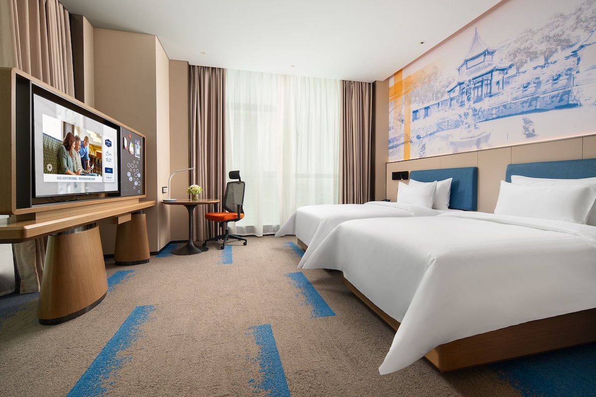 Hotellihuone, jossa on kaksi sänkyä, työpöytä tietokoneella ja suuri seinälle asennettu televisio.