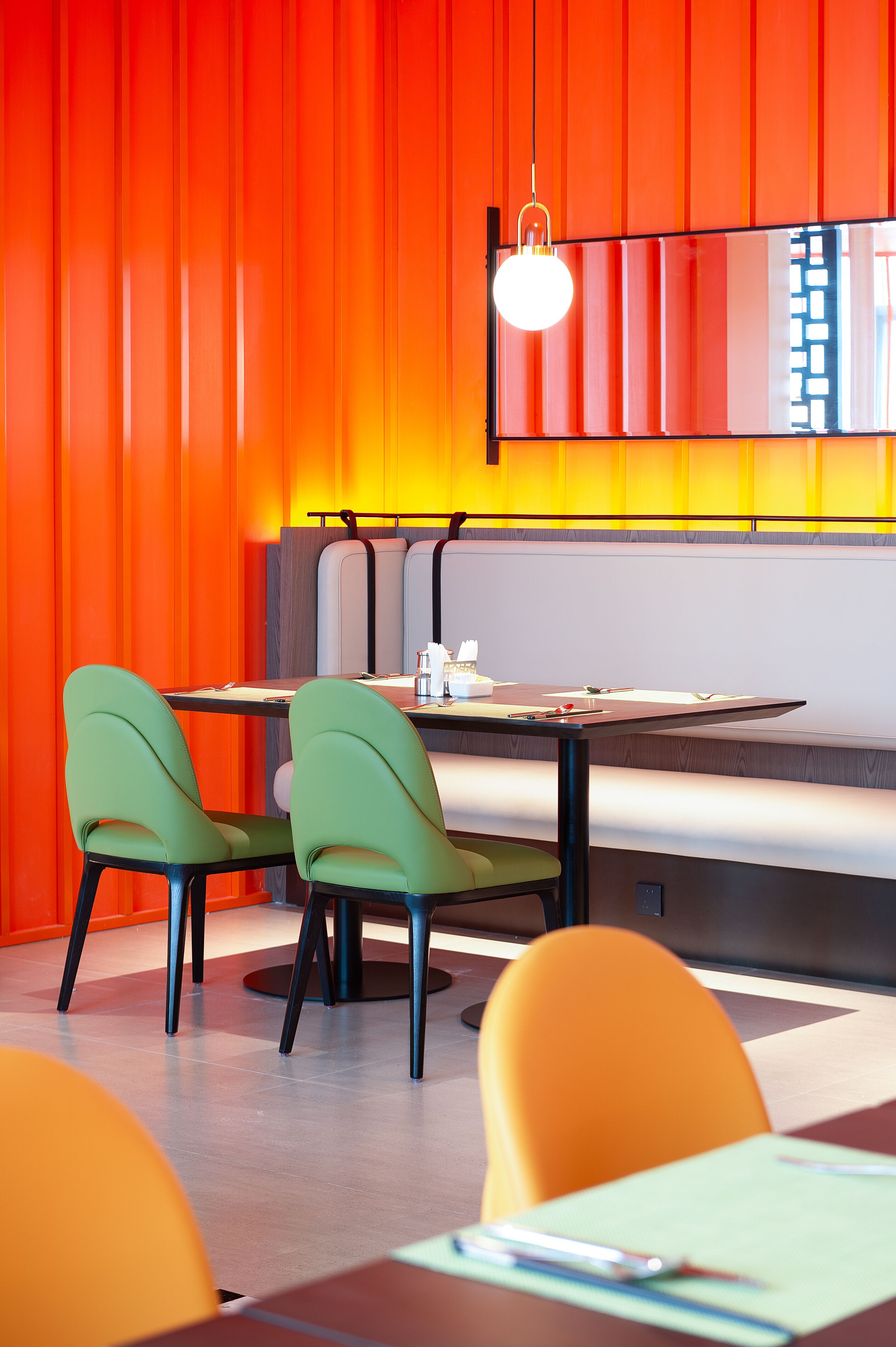 Un restaurant moderne aux couleurs vives, avec une palette de tons orange et jaune, comprenant une longue table conviviale entourée de chaises, d’un banc et d’un luminaire suspendu.
