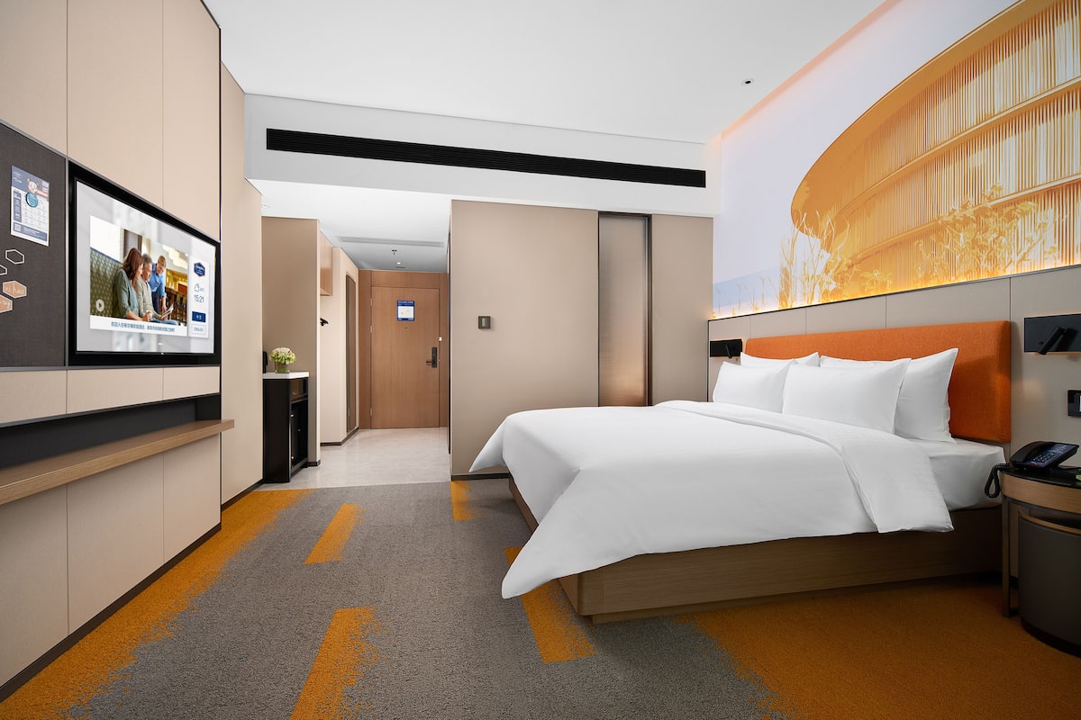 Moderni hotellihuone, jossa on suuri sänky, työpöytä puhelimineen ja seinälle asennettu televisio.