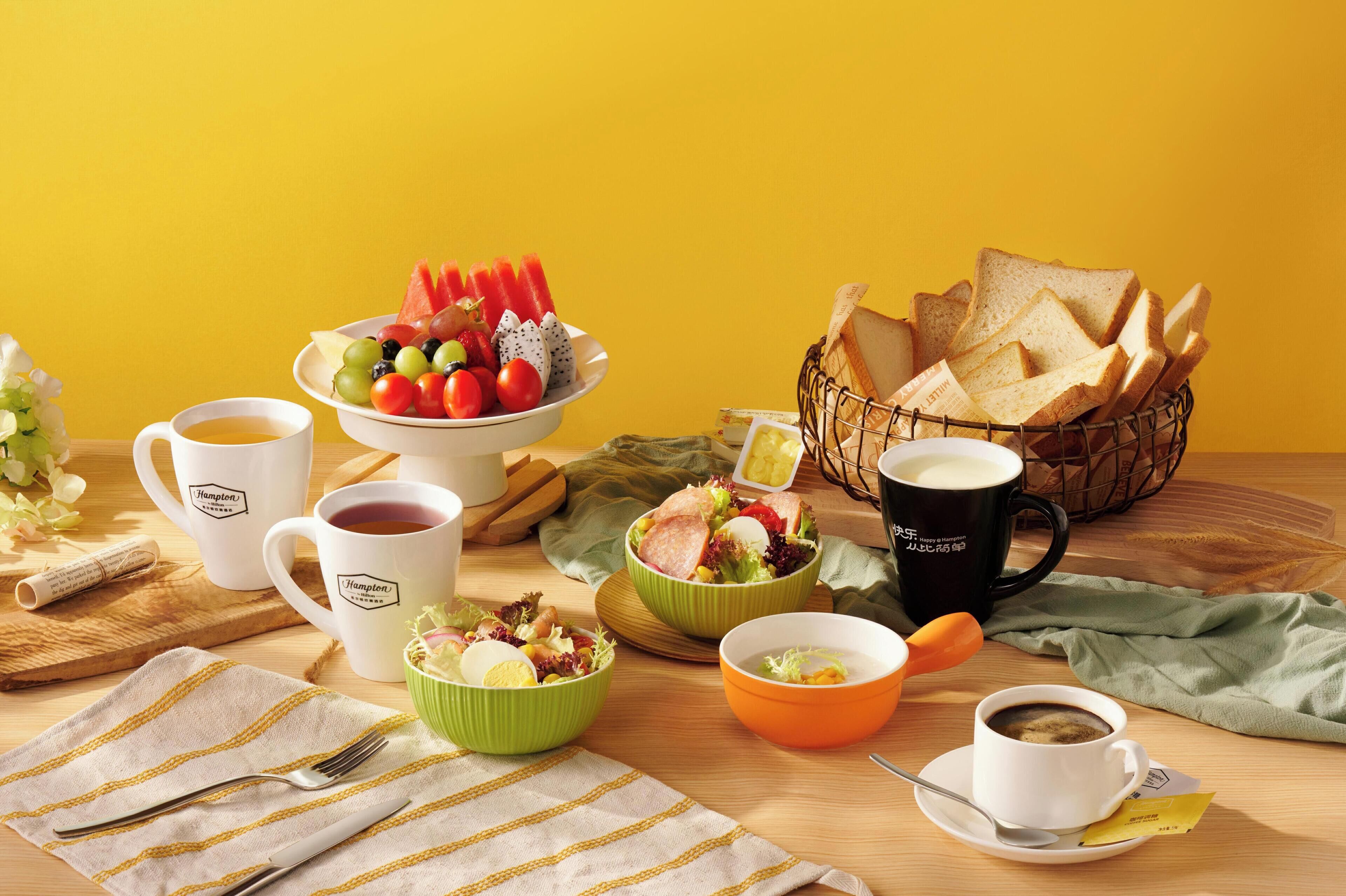 A breakfast setting with various food items and drinks on a wooden table against a yellow background.