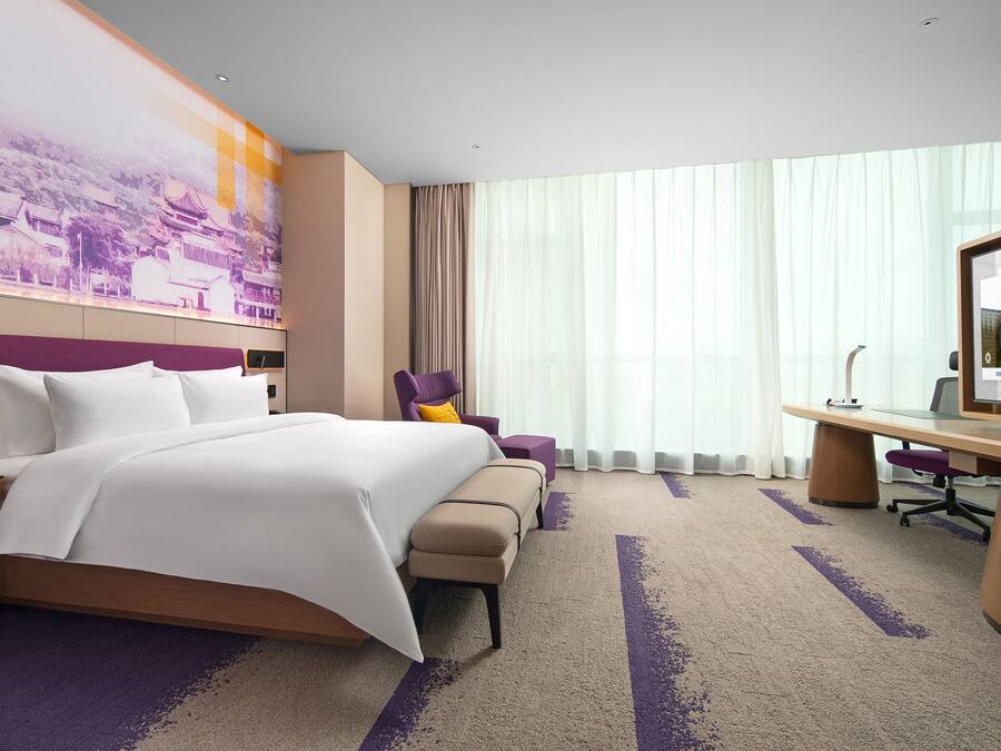 Una habitación de hotel con una cama grande, un escritorio, una silla y un amplio ventanal con cortinas.