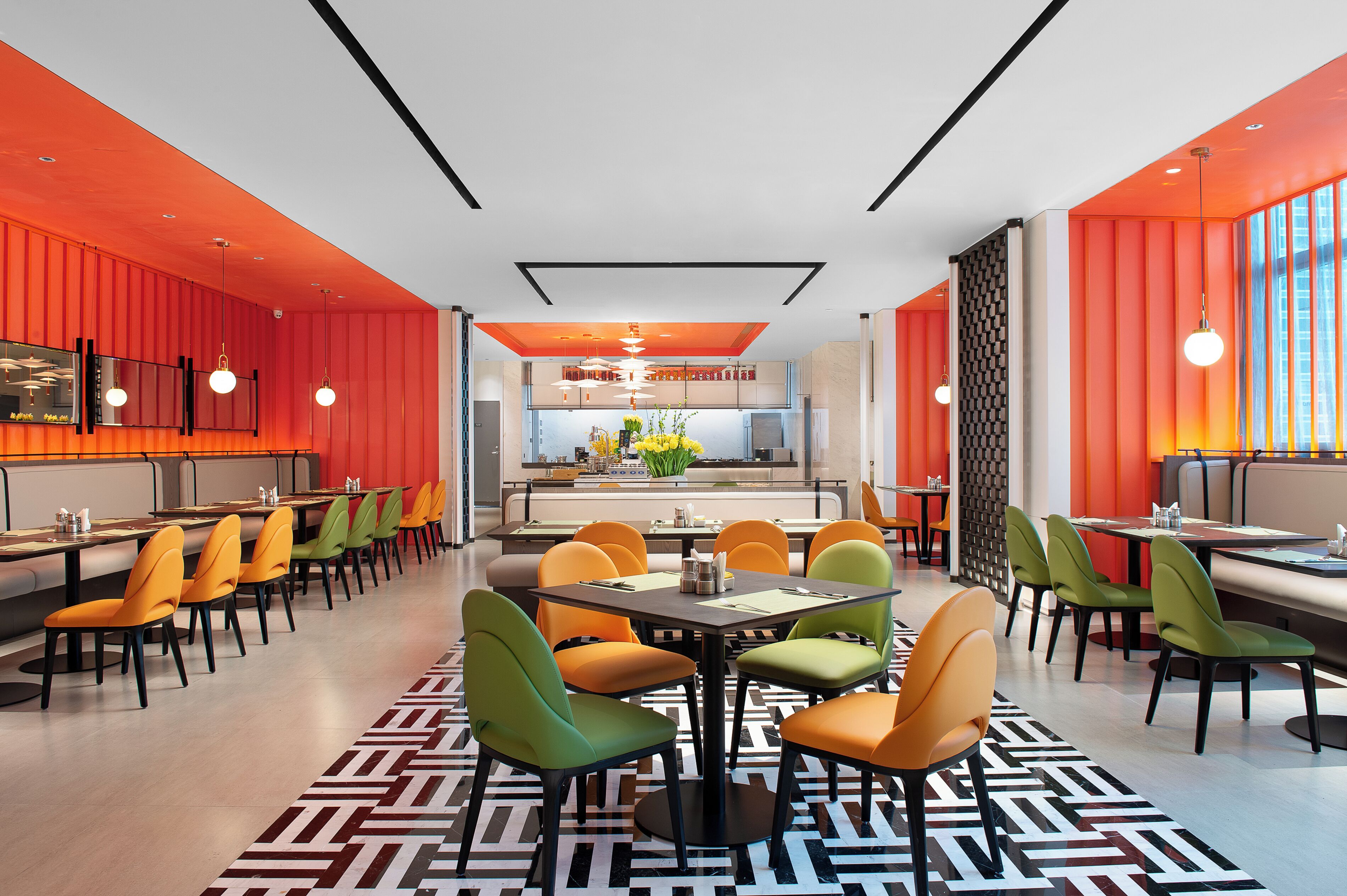 A modern restaurant with vibrant orange walls, a central dining area with colorful chairs, and a visible kitchen in the background.