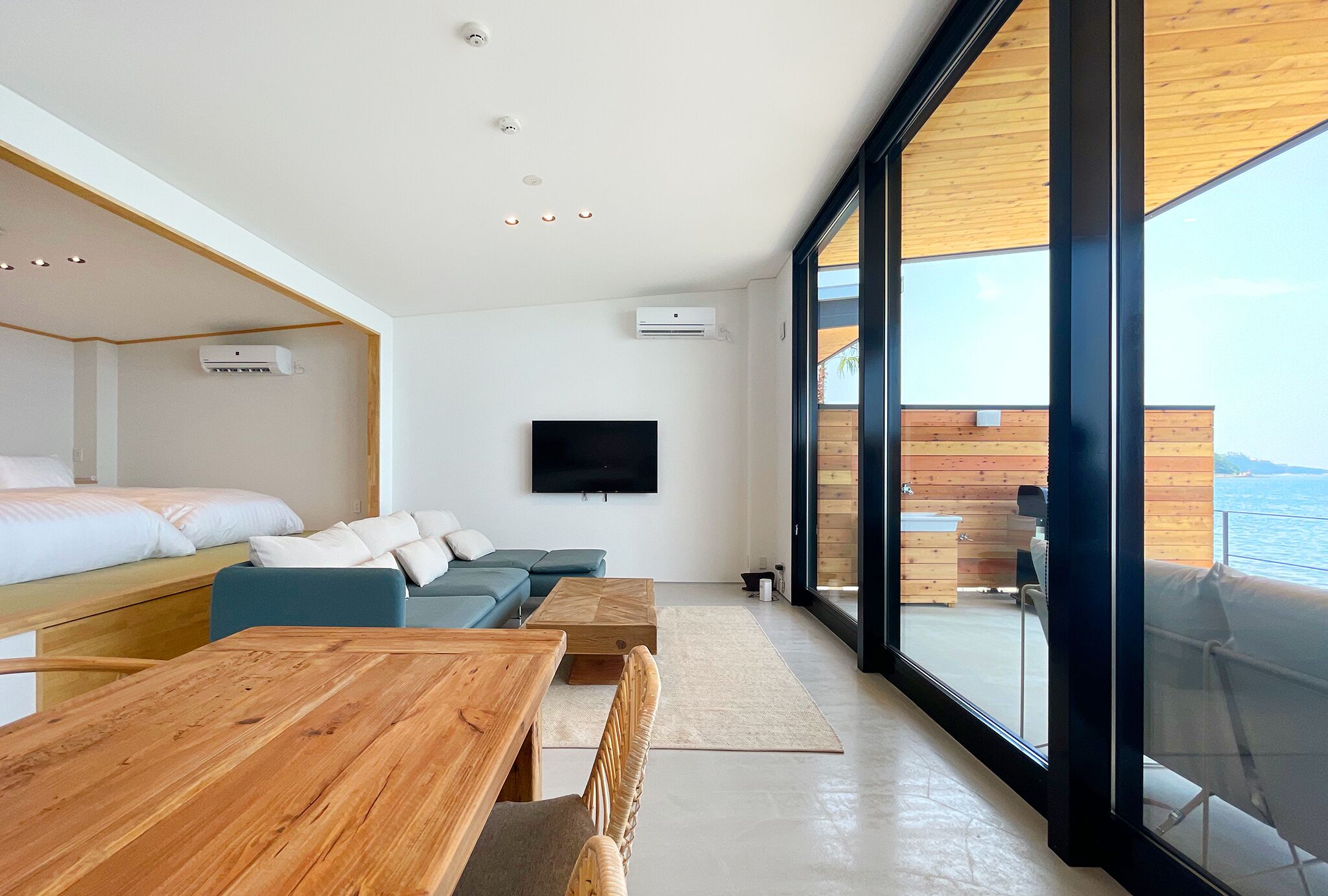 A modern room with a wooden table, a sofa, a TV, and a large window overlooking the sea.