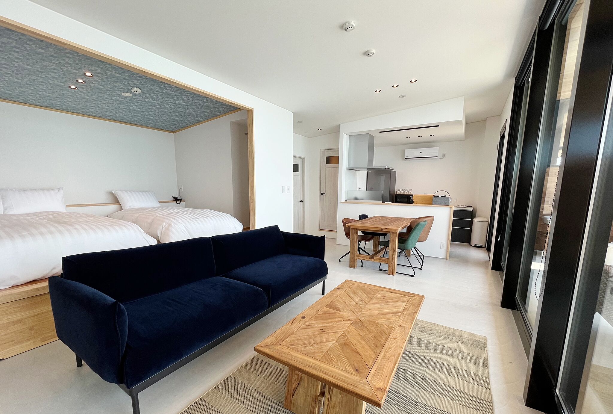 A modern hotel room with a bed, a dark blue sofa, a wooden coffee table, a kitchen area with a dining table and chairs, and large windows.