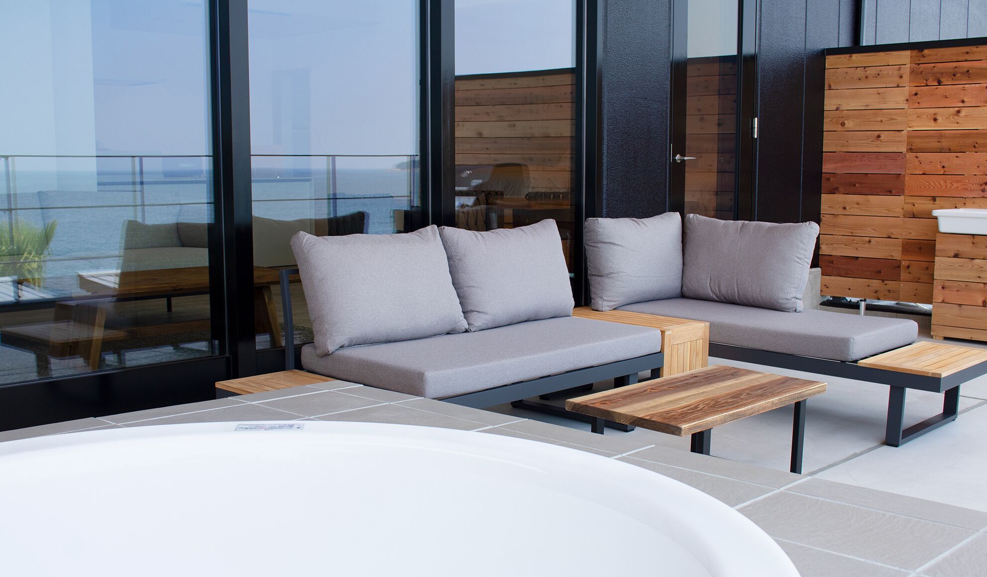 Een modern buitgedeelte met een bank en twee houten tafels, een groot raam met uitzicht op de zee, en een wand van houten panelen.