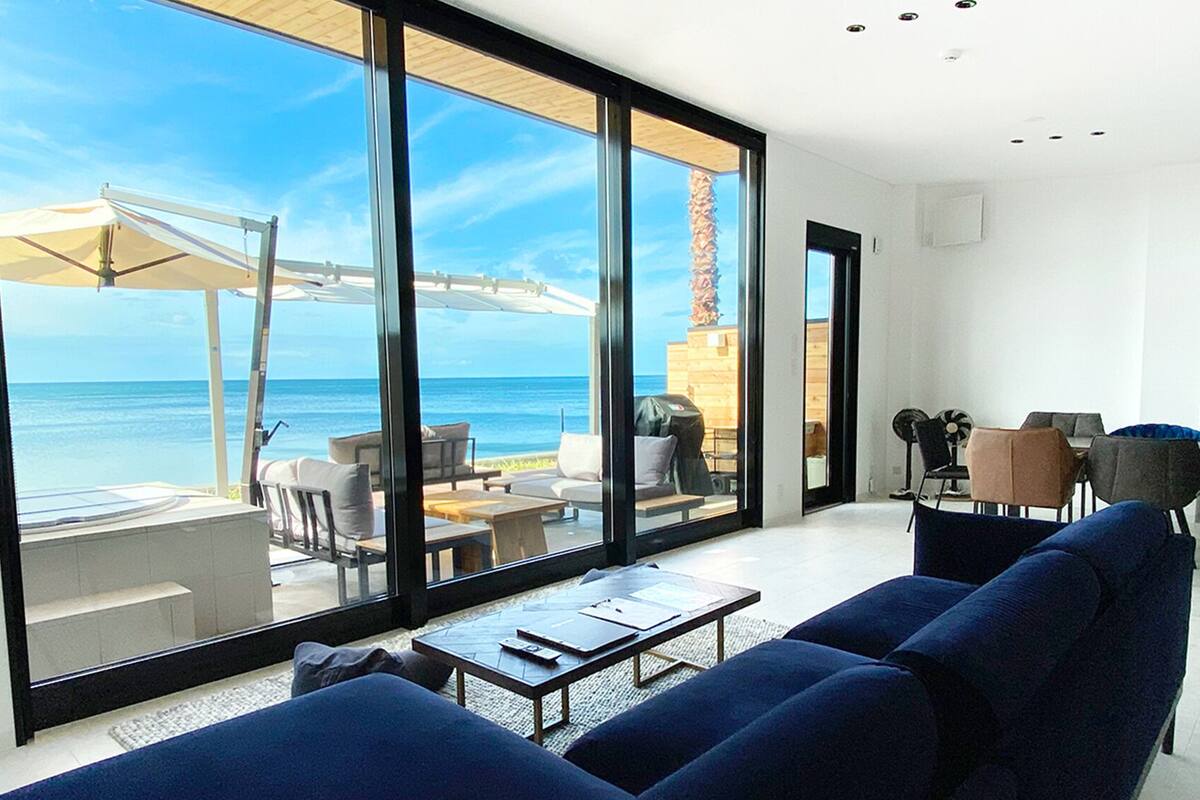 Een moderne woonkamer met een grote glazen wand met uitzicht op zee en een houten terras. Er zijn blauwe banken, een salontafel en een eethoek met stoelen.
