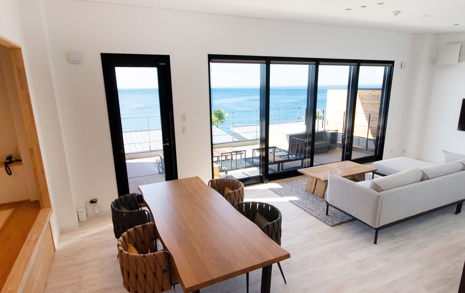 Un soggiorno moderno con un tavolo di legno, due sedie, un divano e un'ampia porta scorrevole in vetro che si apre su un balcone con vista sul mare.