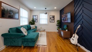 A living room with a green sectional sofa, a small round coffee table, a white guitar stand, and a TV on a wooden wall. - Taylor Swift Eras Inspired Home-10 min to Broadway Sleeps 10 (Nashville)