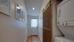 A laundry room with a front-loading washer and dryer, wooden slat door, and framed pictures on the wall. - Taylor Swift Eras Inspired Home-10 min to Broadway Sleeps 10 (Nashville)