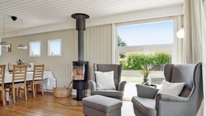 A modern living room with a wood-burning stove, grey armchairs, a dining table with chairs, and large windows offering a view of greenery. - "Volmer" - 700m from the sea by Interhome (Hejls)