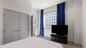 Habitación de hotel con cama, televisor sobre un soporte, armario y ventana con cortinas.