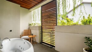 Ein modernes Badezimmer mit freistehender Badewanne, Holzboden und Blick ins Grüne durch ein Fenster mit horizontalen Jalousien.