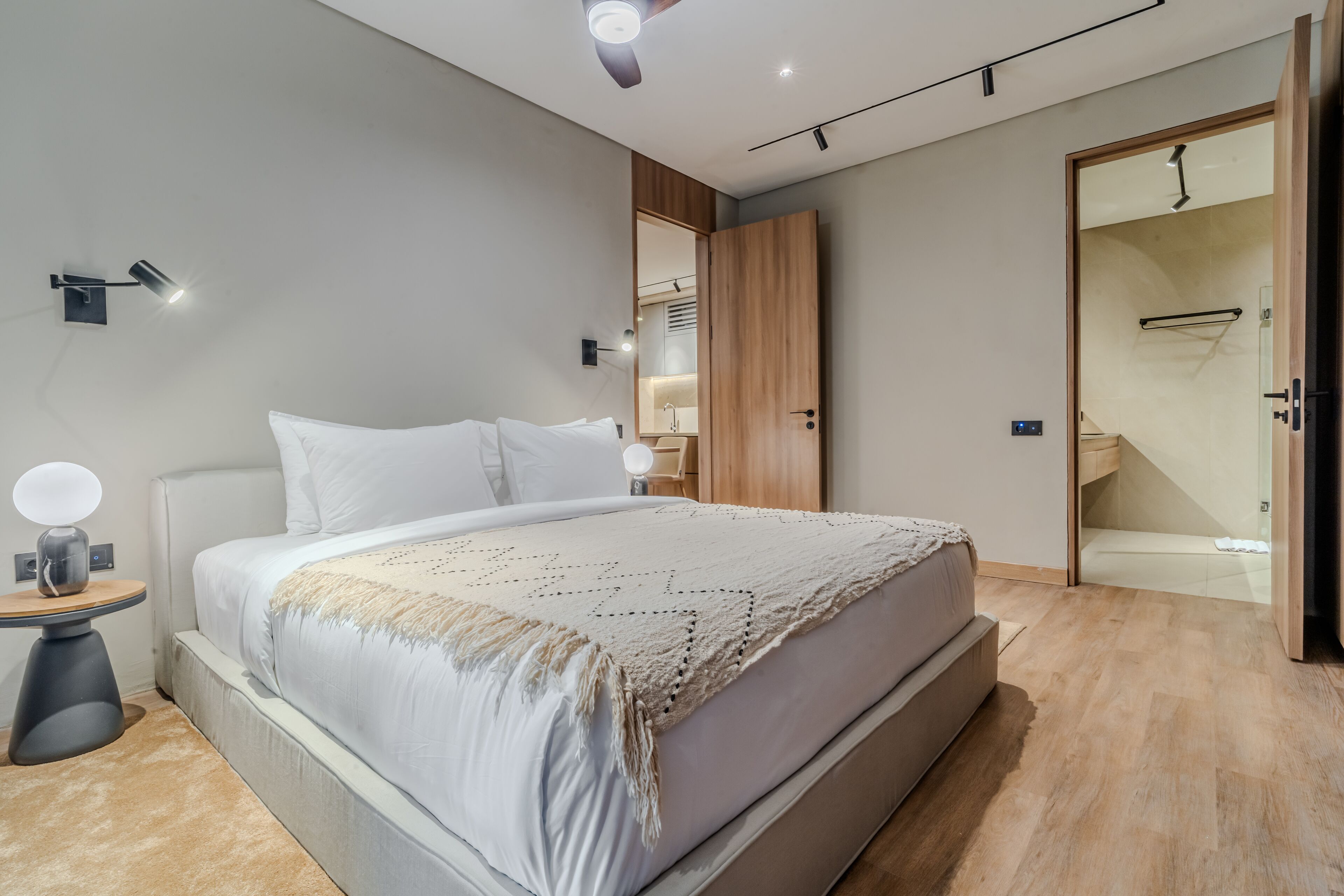 A modern bedroom with a large bed, bedside tables, a nightstand, a wardrobe, and a bathroom visible through an open door.
