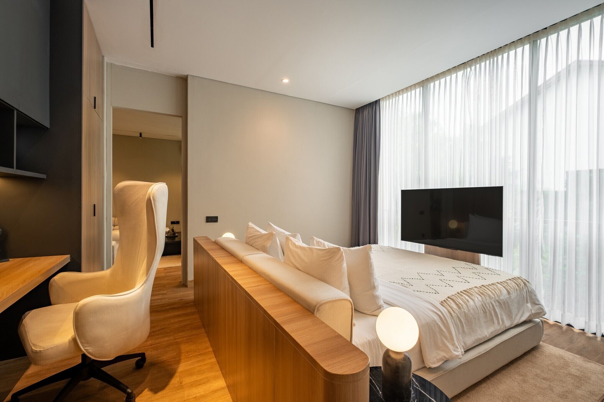 A modern hotel room with a long wooden bench, a white sofa, a TV, and a large window with sheer curtains.
