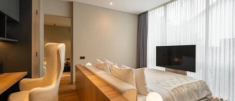 A modern hotel room with a long wooden bench, a white sofa, a TV, and a large window with sheer curtains.